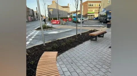 Cumberland Council View of a road junction, with a pavement area foremost bordered by a verge with one a sapling and small plants, with two bench seats, one curved and ne straight. The road in front of it curves round to an open area on which two buses are parked, with buildings behind them and to the left a straight road bordered by terraced houses stretching into the distance.