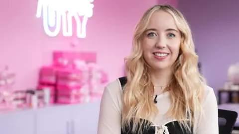 A woman with long blonde hair in a black and beige shop, with a pink-purple blurred background