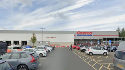 A large Costco warehouse with a car park in front, showing parked cars, people walking and the store entrance.
