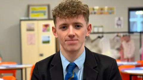 A boy with short curly blond hair is looking into the camera.He is dressed in a school uniform - a dark coloured blazer, a light blue shirt, and a blue and yellow striped tie. The background is out-of-focus, but you can make out a classroom with tables, orange chairs and cabinets. 