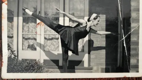 A black and white photo of Joy Saunders during her youth doing a tap dance. She is wearing a black dress and tights and posing with her left leg on the ground and right foot in the air behind her.