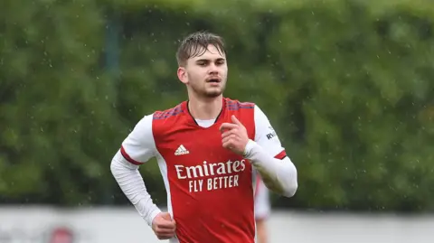 Getty Images Billy Vigar with wet hair, running in the rain. He wears a red arsenal top with white sleeves.