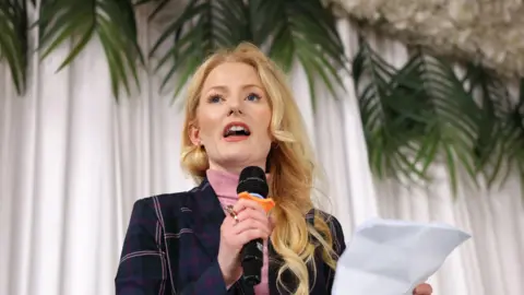 PA Media Hannah Spencer speaks into a microphone at an event. She holds a piece of paper and stands in front of backdrop of a white drape and green plants hanging down.