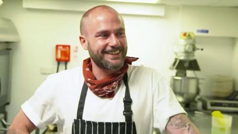 A bearded man - Russ Ball - wearing a bandana around his neck and an apron smiling in a kitchen