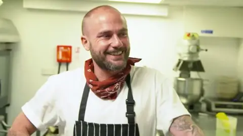 BBC A bearded man - Russ Ball - wearing a bandana around his neck and an apron smiling in a kitchen