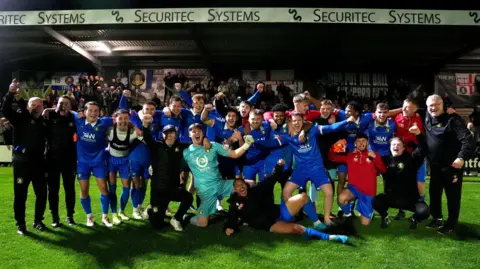 PA Media Football players in blue kits and coaches wearing black celebrate an FA Cup victory on the pitch in front of a stand.