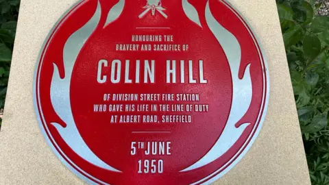 BBC/Adele Forrest A red round plaque with the name of a firefighter who died in 1950 sits on top of a plinth.