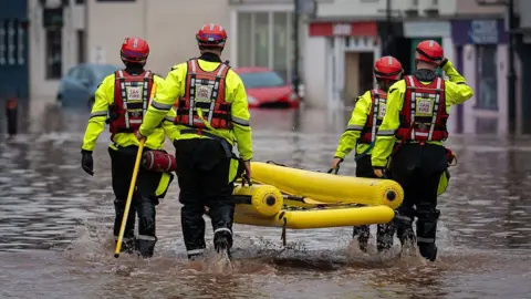 Matthew Horwood/Getty Images Fire service workers walk through water carrying an inflatable dinghy during flooding in Monmouth in November 2025