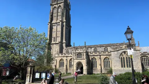 BBC Image of St John the Baptist's Church on a busy high street. There is a blue sky behind the church and in the foreground there is greenery, an iron gate and some people. 