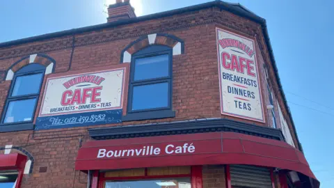 The outside of a cafe with a red canopy that says 'Bournville Cafe' and signs above it on the brick wall that read 'Bournville Cafe' and 'Cafe, Breakfasts, Dinners, Teas'.