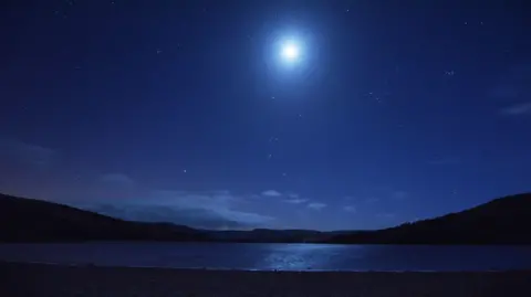 James Allinson The moon shining brightly in the dark, streetlight-free sky over Semerwater, Wensleydale, in the Yorkshire Dales National Park. The sky appears dark blue. Some low cloud is hanging over the hills which are framing the lake.