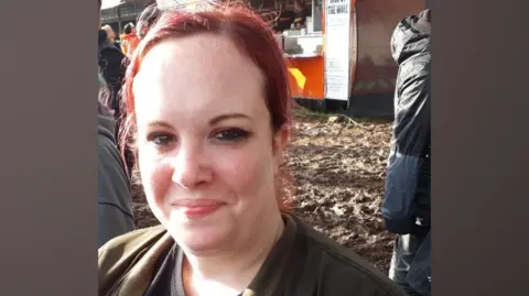 A woman wearing a black top sits down and looks towards the camera with a smile. She has dyed red hair.
