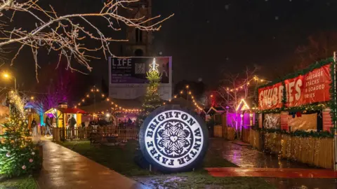 A circular sign which reads Brighton Winter Fayre and with wooden stalls around it lit up with Christmas lights. In the background is a large Christmas tree and a church tower can be seen. It is very wet and the grassy parts of the ground are muddy