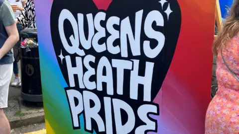 A large colourful piece of artwork spells Queens Heath Pride in white writing on a black heart, with a rainbow background