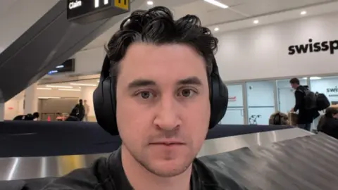 William mengambil selfie sambil mengenakan headphone di depan korsel bagasi di bandara JFK