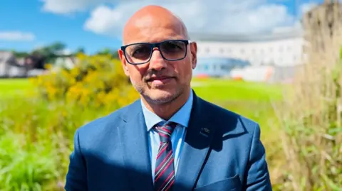Jug Johal, a man wearing a blue suit, smiling, the grounds of a hospital, under a cloudy blue sky.