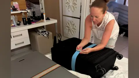 Gavin Sparks A woman wearing a white sleeveless top and with dark red hair that has been tied up unpacks a black suitcase.