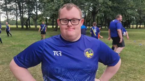 Naj Modak / BBC Jake wearing a blue team shirt and glasses, standing with his hands on his hips on a grassy park field. Other players are training in the background among trees and marker cones.