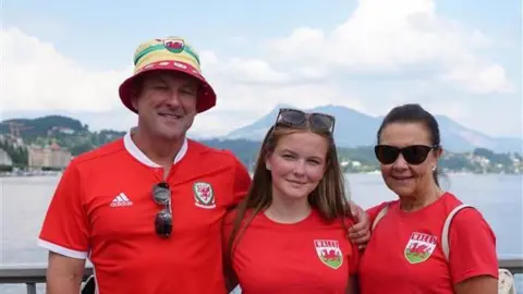 Three people looking at the camera. All have red Wales shirts on and the back drop is of Switzerland's mountains and sea. The man is on the left and has his arm around the girl in the middle, who is his daughter. He has a bucket hat on, in green, red and yellow and sunglasses hanging from his shirt. The girl in the middle has sunglasses on her head and long brown hair. The woman on her right has dark brown hair tied up and sunglasses on.