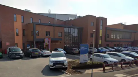 Google outside a hospital, with a car park in front of it. The hospital building center is red brick around the structure with black frames. 