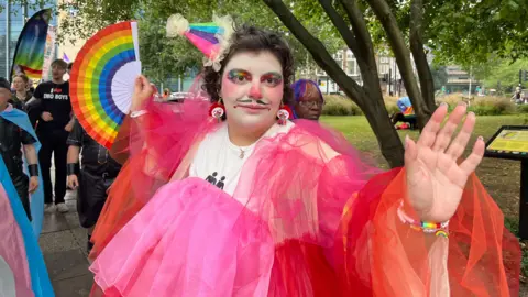 Steve Brock Photography Attendee of the Pride Parade, striking a pose with arms outstretched, wearing a chiffon outfit in shades of pink, red and orange, a rainbow hat at a rakish angle,  and brandishing a rainbow fan.