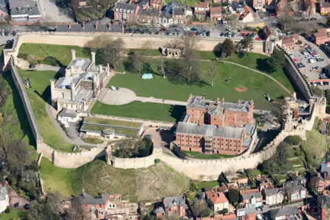 Heritage Images via Getty Images An aerial view of a large castle, with stone walls stretching in an oval shape around large green grounds. Within the grounds stand two large 19th Century buildings, one in stone and one in red brick. Houses and shops can be seen outside the walls.