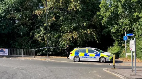 Police car and tape across a wooded area off a roundabout with a sign to Swaythling.