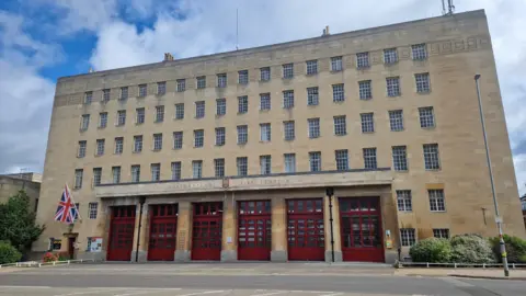 The exterior of the Mounts fire station building in Northampton. It is built from stone blocks and rises over six floors. A Union Jack flag is hanging from one side of the building.