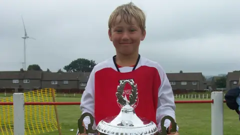 Souttar family John Souttar, when he was a child, is wearing a red and white strip and holding a cup while standing on a football pitch. He is smiling.