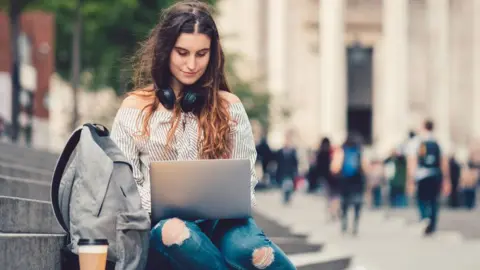 A girl wearing blue jeans and a stripy black and white shirt is sitting on concrete steps on her laptop. She has a bag and a coffee beside her. She is outside an old grey building with people walking past.