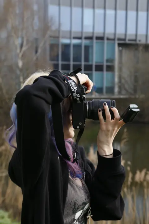Poppy Bellamy Poppy Bellamy - a woman with blonde and purple hair - takes a photo behind a large black camera. She is wearing a blood oxygen monitor on one of her fingers.
