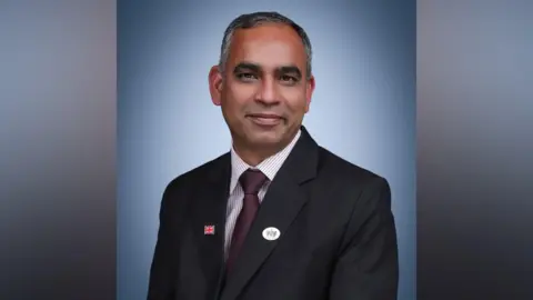 Adi Malviya is wearing a dark-coloured suit with a striped shirt and burgundy red tie. He is smiling and looking at the camera.