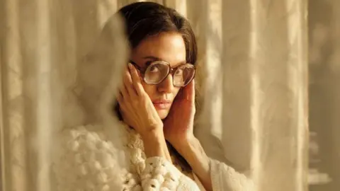 Studio Canal Photo of Angelina Jolie as Callas wearing a white sweater and big glasses