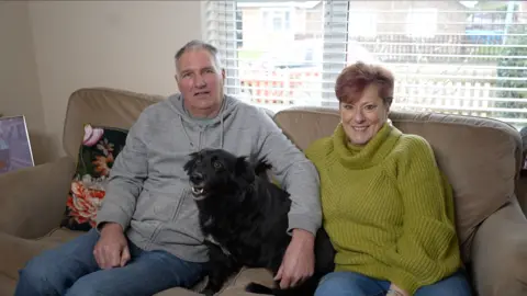 Jamie Niblock/BBC John Hall, wearing blue jeans and a grey hoodie, has his arm around Max, a black dog, which is missing a front left leg, previously amputated. Sitting next to them on the sofa is Lynne, wearing a green roll-neck sweater. She is smiling.