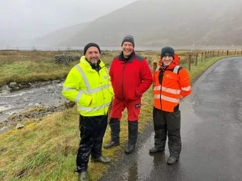 Scottish Water Three men in different coloured waterproof jackets, one yellow, one red and one orange stand next to a small river with a reservoir in the background on a misty day