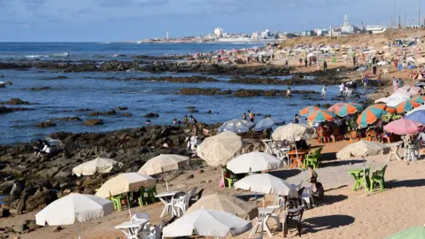 Getty Images Stock image of people sitting under parasol canopies on the sandy beach behing large rocks next to the sea. Casablanca's buildings appear in the backdrop.