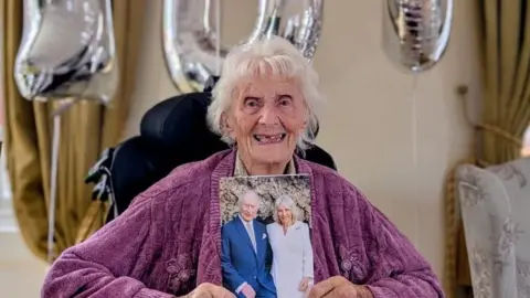 BBC An elderly woman holding up a card with Charles and Camilla on the front. She is smiling.