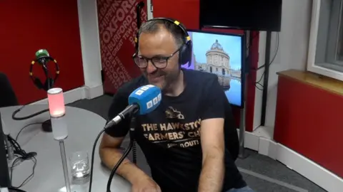 James Florey in BBC Radio Oxford studio, speaking in front of a blue microphone. He is smiling.
