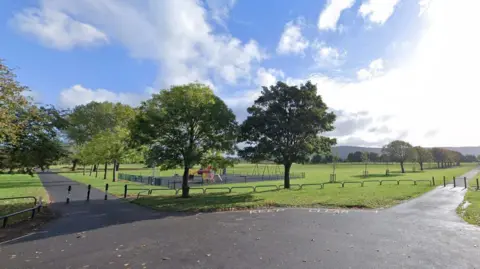 A wide view of a green space in Eston Recreation Ground. Grassy areas are intersected with grey pavement. A small children's play park is seen in behind two trees.