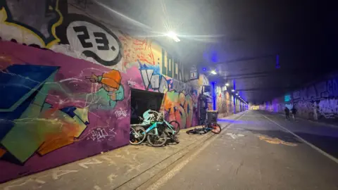 Image shows graffiti covered walls and a pile of abandoned bikes in a former busy underpass that is now a bike highway