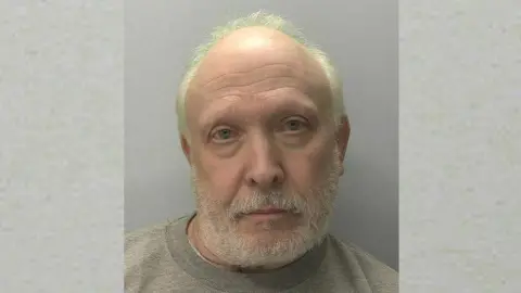 Devon and Cornwall Police Police mugshot of Richard Law. He has white, receding hair and a white beard and he is wearing a grey jumper.