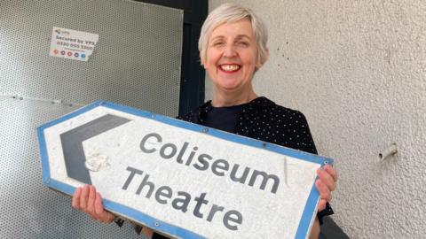 Julie Hesmondhalgh holding up a road sign saying "Coliseum Theatre" in front of the venue's boarded-up main entrance