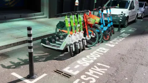 Getty Images A row of rental e-scooters and bikes from Lime, Dott, and other providers is parked in a designated bay marked “E-scooter & cycle hire only” on a city street next to a white van.