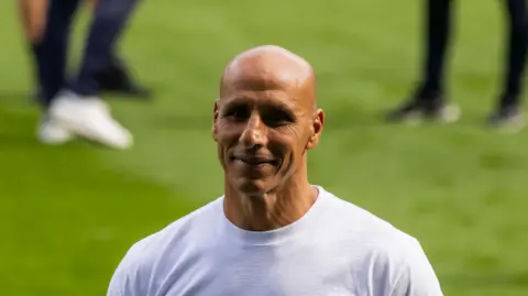 Dino Maamria wearing a white t-shirt standing on a pitch
