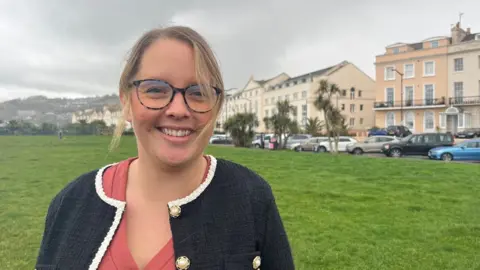 BBC Emma Foreman wearing glasses and a coral top with a blue cardigan with white piping and large gold buttons. She is standing on the green in Teignmouth.