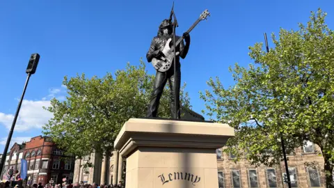 BBC A black-coloured statue of Lemmy holding a bass guitar and singing up towards a microphone. It's been placed on a plinth with "Lemmy" on it. Two trees are next to the statue with buildings visible in the background.