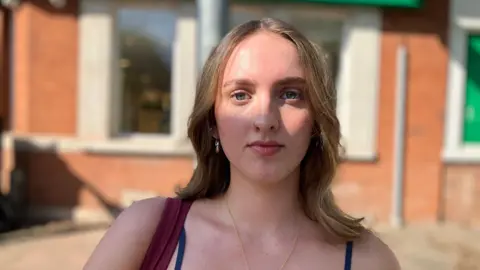 A student in the Leeds suburb of Headingley. She has brown hair and a red brick building is blurred behind her.