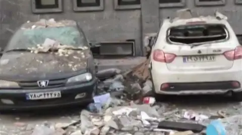 Two cars, a dark coloured Peugeot and a white hatchback both sit on the road in front of the building where the blast happened, covered in rubble