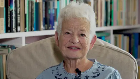 Jacqueline Wilson looks at the camera, sat on an armchair. She is wearing a blue blouse and is sat infront of a bookcase
