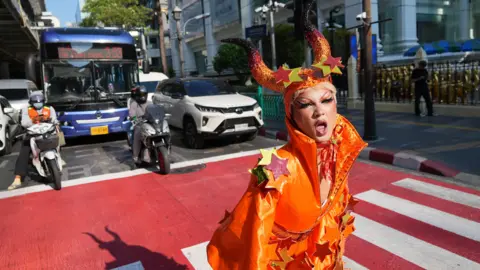 Gawdland, Thai winner of Ru Paul's Drag Race UK versus the World, standing on a red and white crosswalk in Bangkok, with two motorcycles, cars, and a big blue bus behind her, stopping for a red traffic light